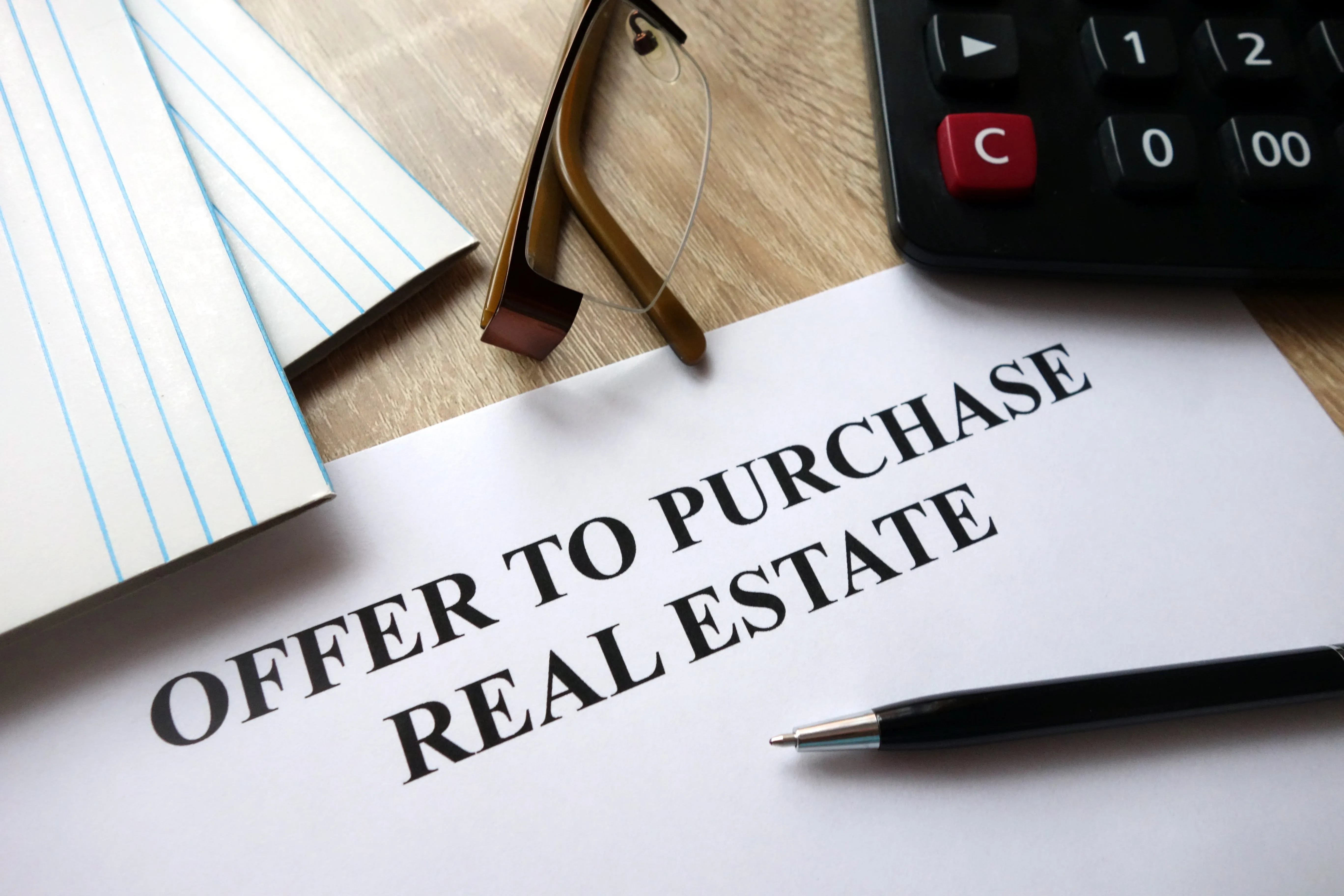 Papers, calculator, pen and a pair of glasses next to documents that say offer to purchase real estate. 