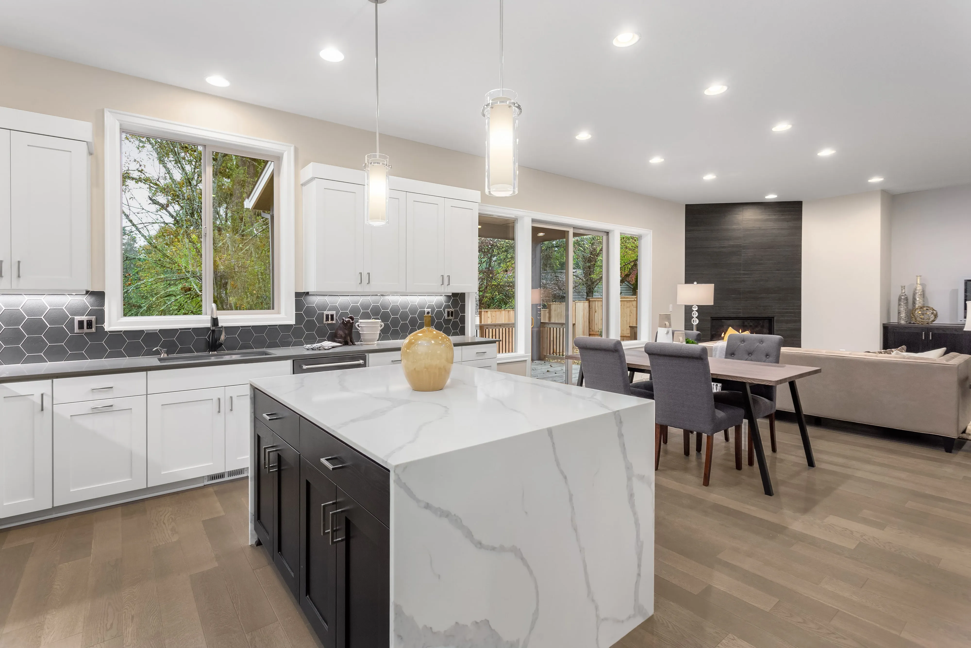 A photo of a kitchen in a luxury home. The counters are marble with a black pentagon tiles backsplash. The cabinets are white. You can see the living room with a nice fireplace with black tile. 