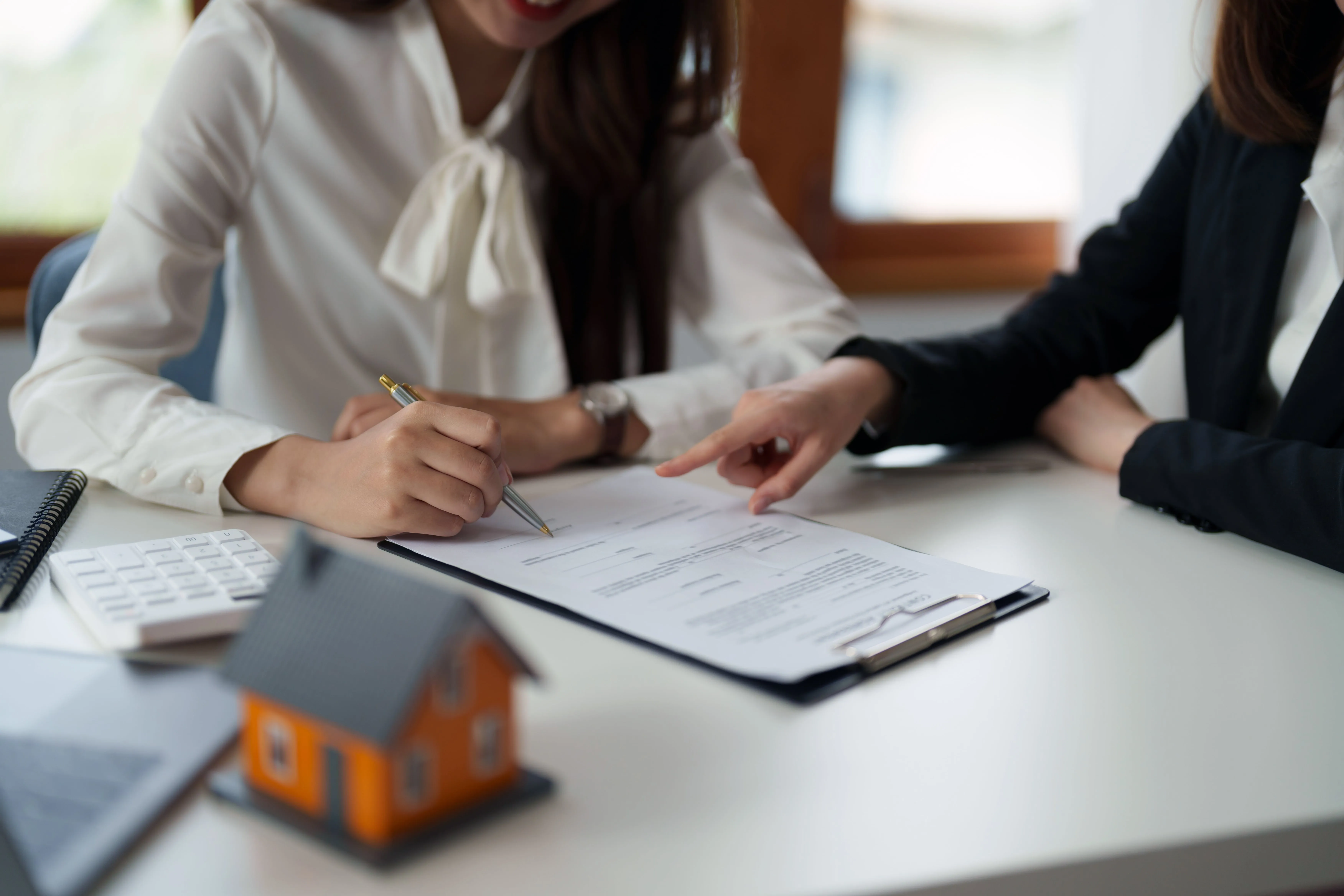 A woman is giving guidance to another woman that is signing papers to complete a real estate transaction.